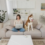 A woman sitting with a laptop on her knees beside her dog