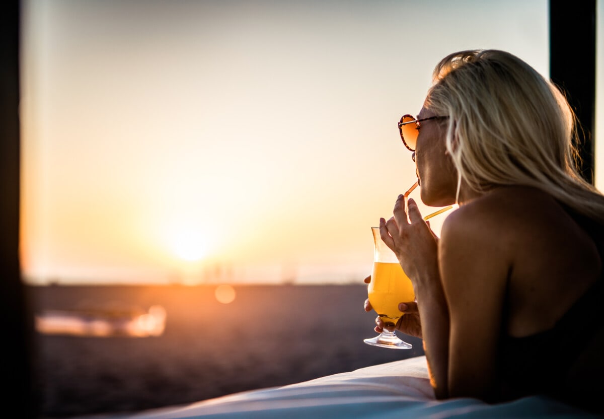 Young woman drinking summer cocktail on the beach bed at sunset.