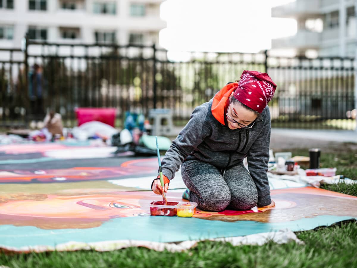  woman creating outdoor mural on the lawn. She is dressed in casual outfit, wearing bandana and eyeglasses. Exterior of public park in the city.