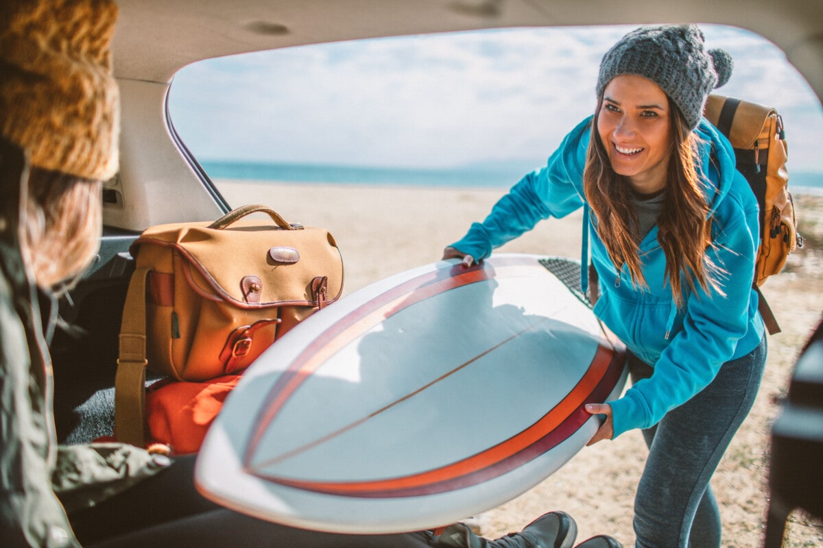Photo of a surfer girl packing for surfing road trip: putting a surfboard in car trunk