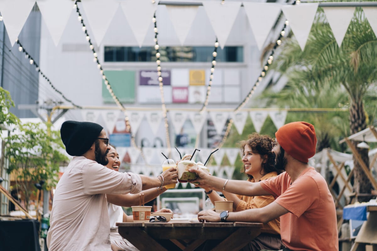 Group of friends making a toast with a coconut drink. They are sitting at a table in the food street market, having fun together.