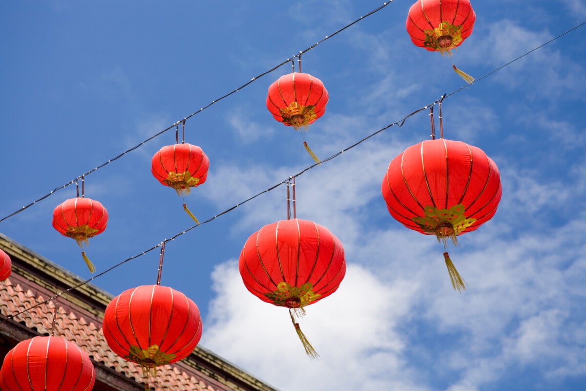 Two diagonal rows of sunlit red Chinese lanterns hanging above a rooftop, against a blue sky