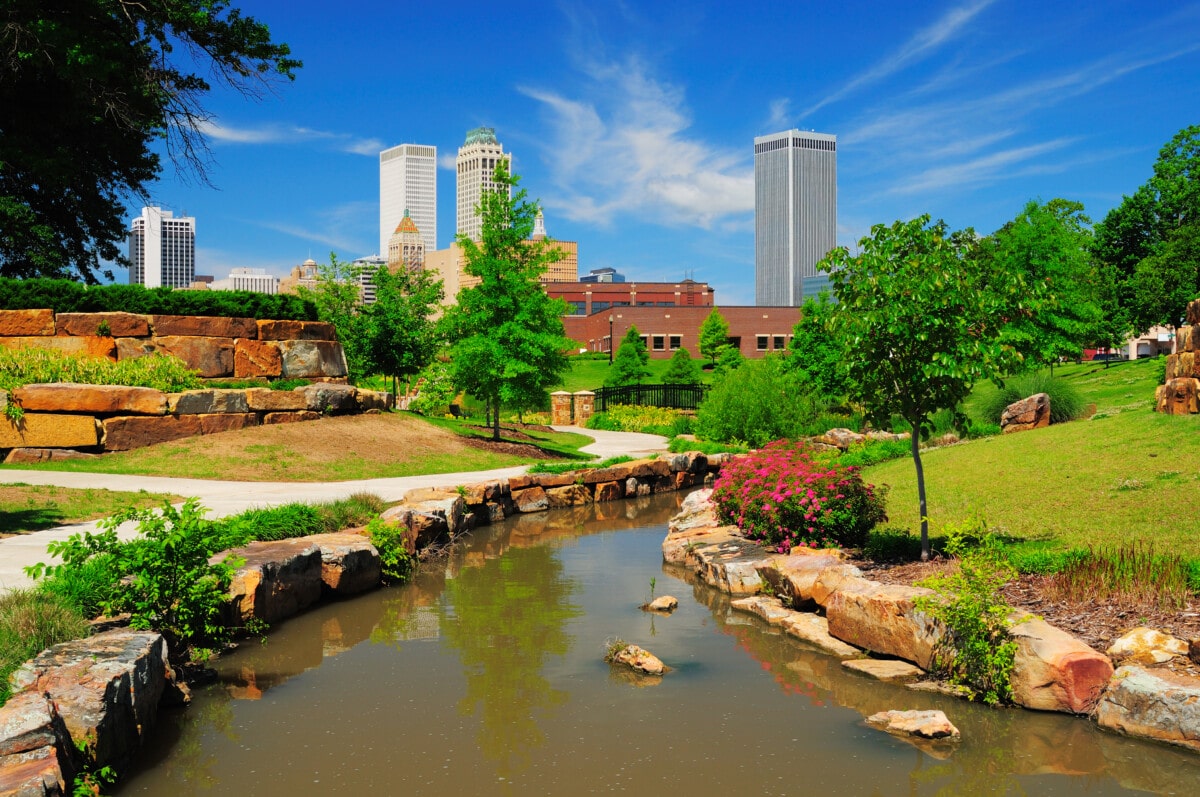 Tulsa downtown skyline from a park with trees, grass, rocks, and a stream in the foreground.