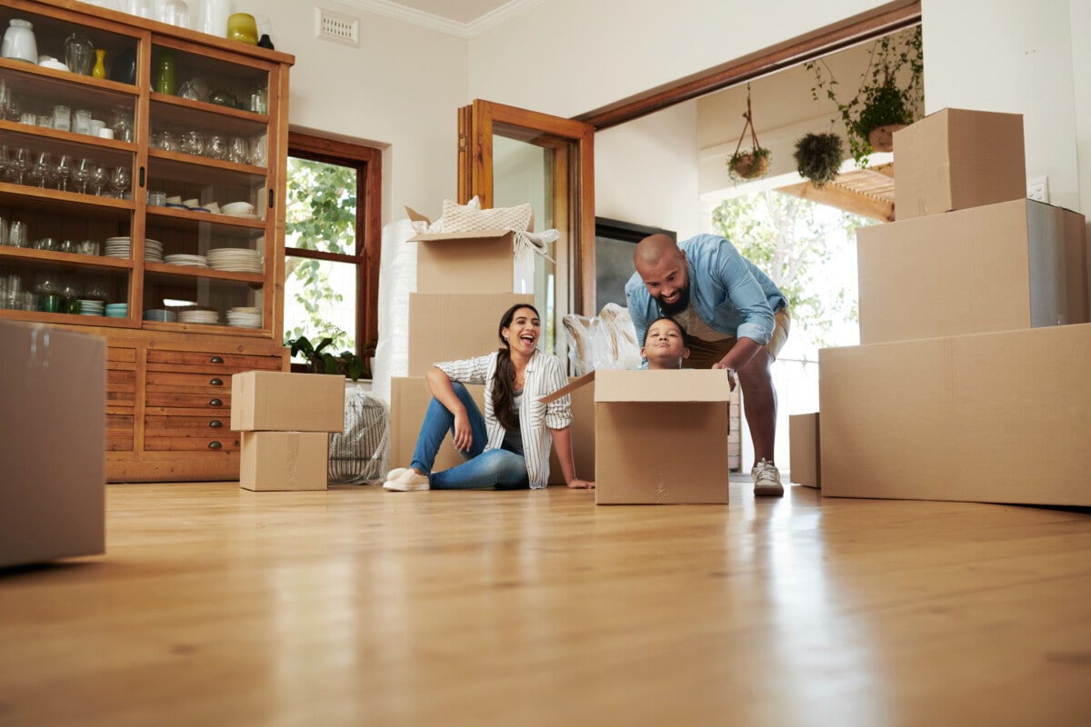 hot of a husband pushing his toddler in a moving box while the wife relaxes in the background in their new home-getty