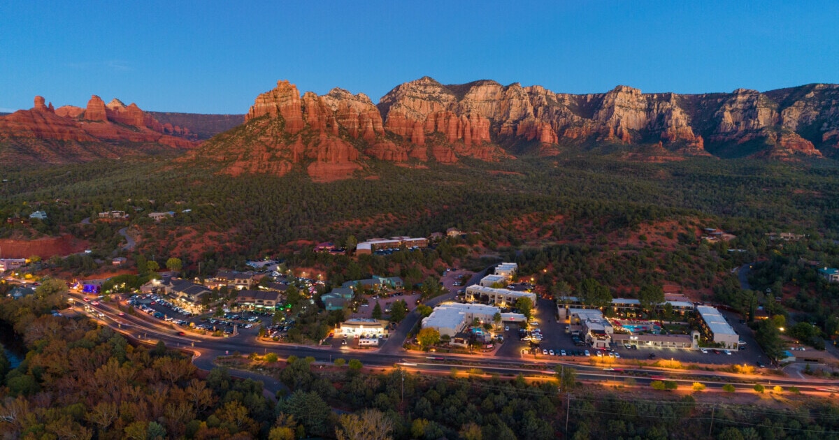 Sedona Verde Valley aerial at dusk during autumn, with mountains in the background and a highway with stores and forests in the foreground.