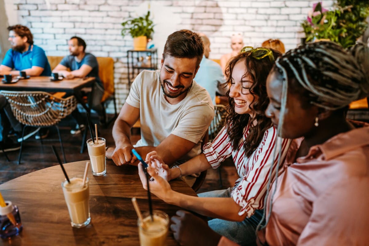 Young group of friends relaxing in a café, drinking juice and coffee and scrolling through social media using smart phone.