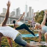 Group of people attending to a yoga class outdoors at sunset with New York cityscape on their background. They are meditating and relaxing.