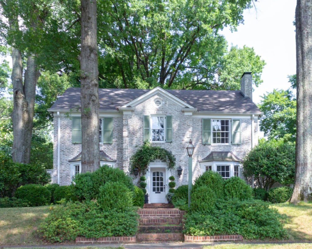 white large brick home with green shutters surrounded by large green trees