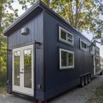 A dark blue granny pod, a type of tiny home, is parked on a gravel path surrounded by trees. The granny pod has a sloped roof, white-framed windows of varying sizes, and double white doors at the end.