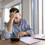 Worried tired homeowner getting financial problems, using calculator at home office workplace to calculate mortgage, touching head in frustration, finding bankruptcy risk, making difficult decision