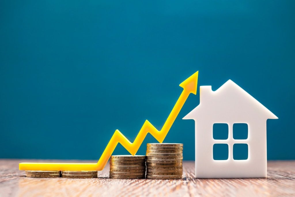 A white, cutout-style house sits on a wooden surface next to stacks of gold coins that gradually increase in height, representing growing equity or value. A bright yellow arrow dramatically curves upwards from the lowest stack of coins, pointing towards the top right, symbolizing an increase. The background is a solid, deep teal color. This image visually represents the concept of increasing home equity and financial growth, which is often a consideration when asking "Do you need a down payment to refinance?"