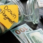 A glass jar, labeled with a yellow sticky note shaped like a house and the words "Down Payment," is tipped on its side, with money spilling out. Several $100 bills are visible outside the jar, alongside a calculator and more scattered currency in the background, all on a dark wooden surface. This image prompts the question, "Is a Large Down Payment on a House Always Best?"