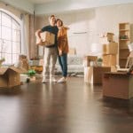 A smiling couple embraces in their new home, surrounded by moving boxes, a ladder, and houseplants. They are standing on a polished wood floor in a sunlit room with an arched window, capturing a happy moment during the process of moving in.non contingent offer