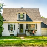 An older woman tends to flowers in front of a small white house on a green lawn surrounded by trees. If someone inherits her home, they might want to use an ibuyer or cash-offer company to sell it.
