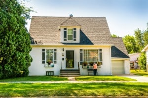 An older woman tends to flowers in front of a small white house on a green lawn surrounded by trees. If someone inherits her home, they might want to use an ibuyer or cash-offer company to sell it.