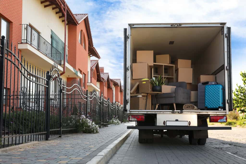 A moving truck is parked outside of a row of townhomes. The back is open and shows the truck packed with boxes, furniture, and specialty items.