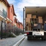 A moving truck is parked outside of a row of townhomes. The back is open and shows the truck packed with boxes, furniture, and specialty items.