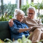An older man and woman sit together on a couch looking at common reasons why people move to retirement communities.
