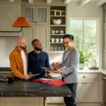 Male real estate agent stands with two male customers in a kitchen